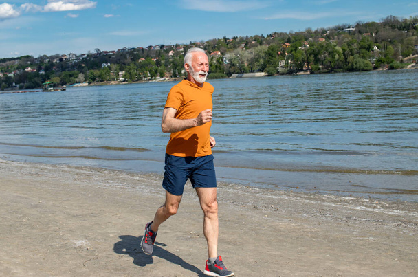 Mutlu, enerjik emekli adam kumsalda koşmaktan zevk alıyor. Nehir kenarında koşuyor.. - Fotoğraf, Görsel