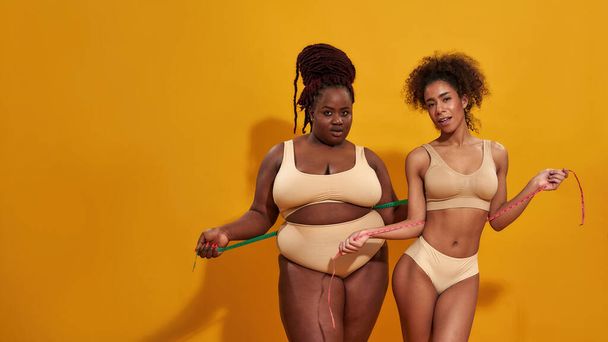 Two african american women in underwear with different body shape holding metric tape measure, standing together isolated over orange background - Foto, Imagen