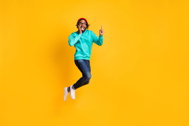 Full size profile photo of brunette optimistic guy jump point empty space wear cap spectacles pullover jeans isolated on yellow background - Fotoğraf, Görsel