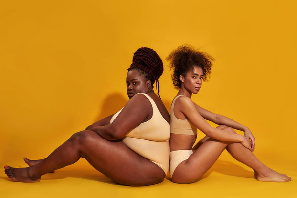 Full length shot of two diverse african american women in underwear with different body size sitting back to back isolated over yellow background - Fotografie, Obrázek