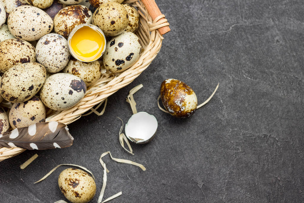 Quail eggs and broken egg in wicker basket. Egg and eggshell on table. Black background. Top view. Copy space - Zdjęcie, obraz