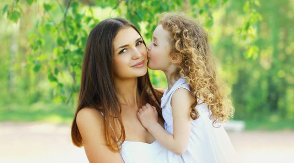 Portrait of little child daughter kissing her happy smiling mother on the grass in a summer day - Φωτογραφία, εικόνα
