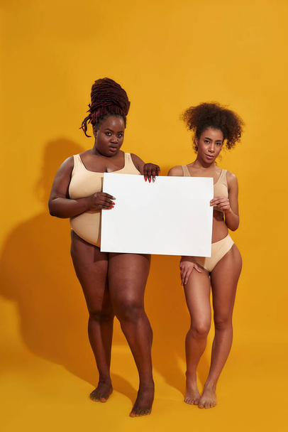 Full length shot of two confident african american women in underwear with different body weight looking at camera, holding blank banner, standing isolated over yellow background - Valokuva, kuva