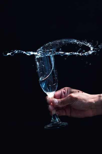 View of a glass goblet filled with water shaken by hand with black background - Фото, изображение