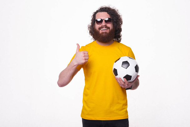 Brodaty młody hipster uśmiecha się ładnie do kamery w okularach przeciwsłonecznych i trzyma piłkę nożną pokazuje kciuk w górę , - Zdjęcie, obraz