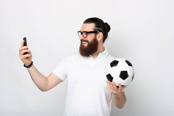 Γενειοφόρος νεαρός άνδρας έχει μια κλήση με κάποιον στο τηλέφωνό του και χαμογελώντας κρατά μια μπάλα ποδοσφαίρου κοντά σε ένα λευκό τοίχο . - Φωτογραφία, εικόνα