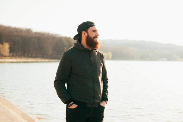 A nice photo of a young bearded man smiling and looking away is standing near a lake . - Φωτογραφία, εικόνα