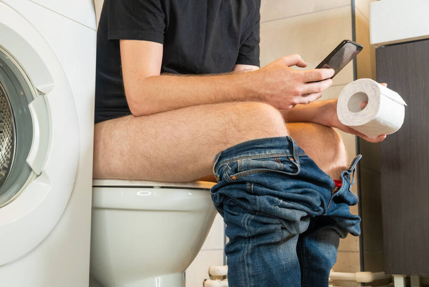 Man sitting on toilet sit bowl with toilet paper and use smartphone, man suffering from diarrhea on toilet bowl at home. - Zdjęcie, obraz
