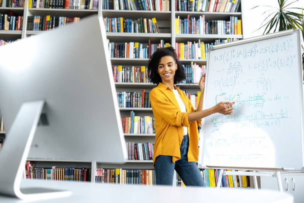 Stilvolle afroamerikanische Trainerin, steht am Whiteboard, in lässiger Kleidung, nutzt Computer, leitet eine Online-Lektion, kommuniziert mit Studenten per Videokonferenz, blickt auf den Bildschirm, Fernunterricht - Foto, Bild