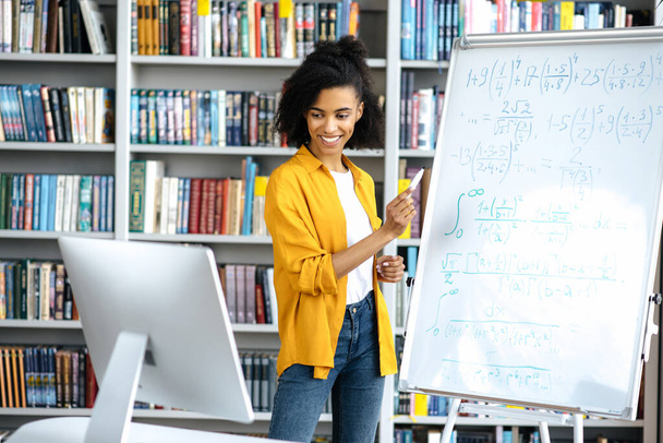 Молодая привлекательная умная афроамериканка, учительница, стоит у доски, пользуется компьютером, проводит онлайн-семинар, общается со студентами по видеосвязи, жестикулирует рукой, улыбается - Фото, изображение