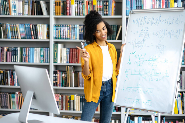 Happy smart selbstbewusst schöne afroamerikanische Lehrerin in stilvoller Kleidung, steht in der Nähe des Whiteboards, leitet eine Online-Lektion, kommuniziert mit den Schülern über eine Videokonferenz, per Computer - Foto, Bild