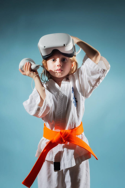 Studio Portrait of young caucasian girl in karate uniform takes off virtual reality helmet after finishing workout or playing video games with virtual reality headset on blue background. - Photo, Image