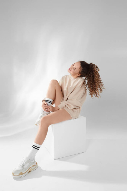 Beautiful model girl with an afro hairdo in a trendy sweatshirt and modish sneakers posing over white background in studio - Foto, afbeelding