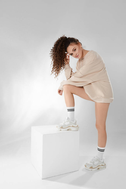 Attractive young model woman in a grey sweatshirt and trendy sneakers posing on white background, studio portrait. Mock up with copy space. Looking at camera - Fotografie, Obrázek