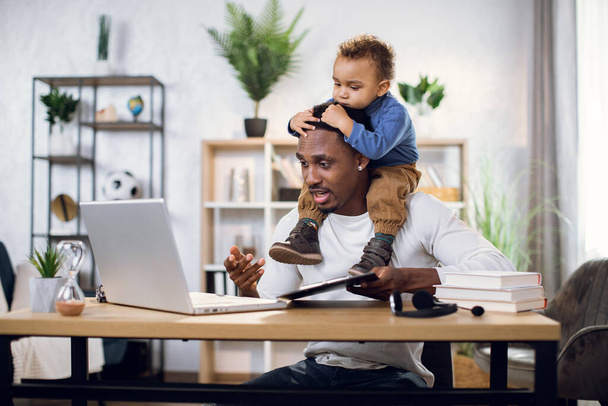 Busy afro man working on laptop with son on neck - Foto, Imagem