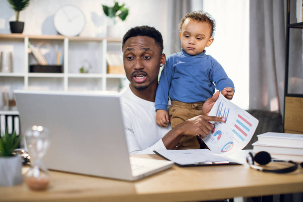 Afrikai üzletember, aki otthonról dolgozik, a fiával a kezében. - Fotó, kép