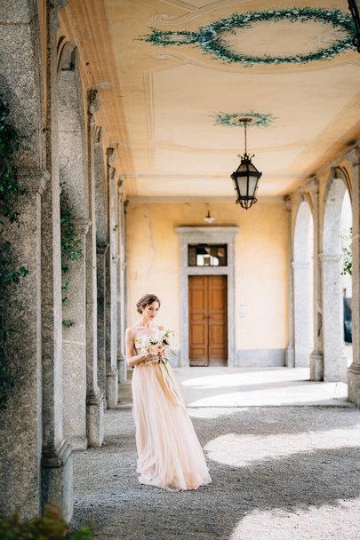 Bride in a beautiful dress with a bouquet of pink flowers stands in an arched hall. Lake Como, Italy - Photo, Image