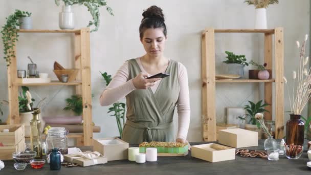 Střední záběr mladé atraktivní ženy fotografování krásného ručně vyrobeného oleje zdobené suché květinové lístky na smartphone - Záběry, video