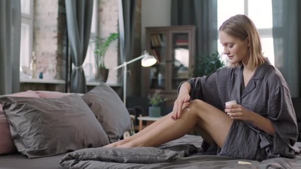Keskipitkä PAN laukaus kaunis nuori valkoihoinen nainen nauttii prosessi soveltaa luonnon orgaaninen vartalovoide hänen hoikka jalat, jotta ne sileä ja kosteutettu istuu mukava sänky silkki kylpytakki - Materiaali, video