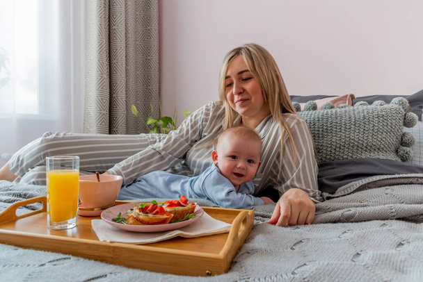 Young woman comfortable gray pajamas with an infant having breakfast while lying the bed in the bedroom. Concept wellness at home. Emotional health young woman - Photo, Image