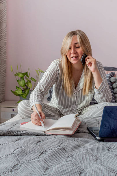Young woman in comfortable gray pajamas works on a computer and talks a cell phone while lying the bed in the bedroom. Business woman maintains a social distance works remotely. New normal concept - Photo, Image