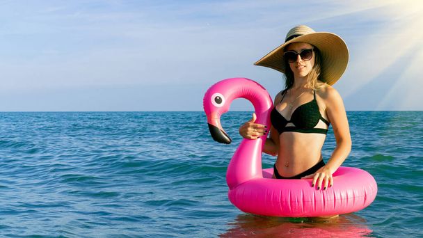 L'heure d'été. Jeune femme sexy en chapeau de paille, maillot de bain bikini, lunettes de soleil avec flamant rose gonflable dans l'eau bleue de l'océan. L'eau de baignade, s'amuser et profiter des vacances de voyage - Photo, image