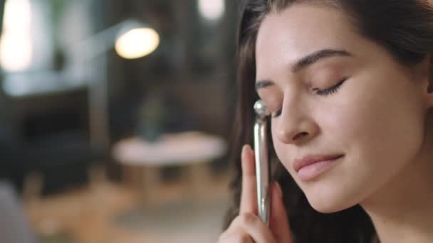 Közelkép fiatal vonzó nő siklás hűvös alumínium súlyzó alakú görgő az ő tiszta bőr élvezi arcmasszázs során a reggeli szépség rutin - Felvétel, videó