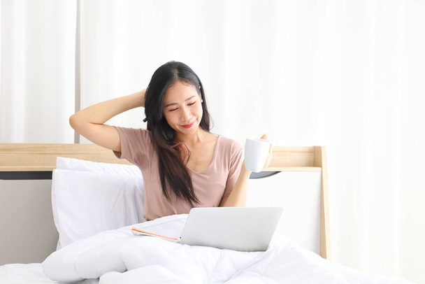 Mladý pěkný asijské dívka pracuje z domova na posteli pomocí počítače notebook pro přístup k její práci on-line s kopírovat prostor - Fotografie, Obrázek