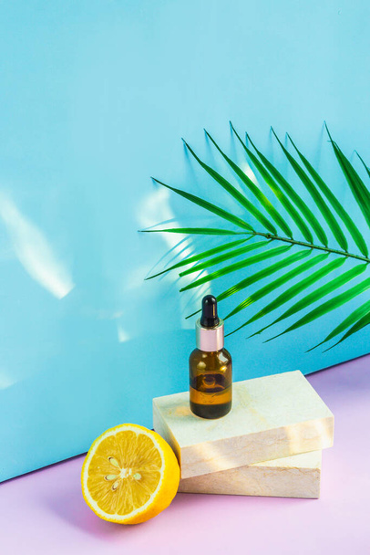 A cosmetic jar with an eyedropper stands on two granite slabs, like on a catwalk, next to half a yellow lemon, a palm leaf and the glare of the sun in the background. Cosmetics for summer. - Photo, image