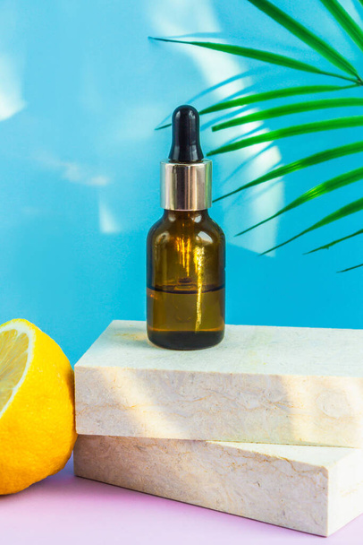 A cosmetic jar with an eyedropper stands on two granite slabs, like on a catwalk, next to half a yellow lemon, a palm leaf and the glare of the sun in the background. Cosmetics for summer. - Foto, afbeelding