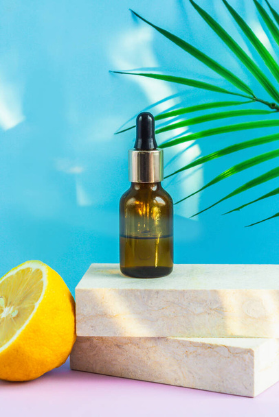 A cosmetic jar with an eyedropper stands on two granite slabs, like on a catwalk, next to half a yellow lemon, a palm leaf and the glare of the sun in the background. Cosmetics for summer. - Photo, Image