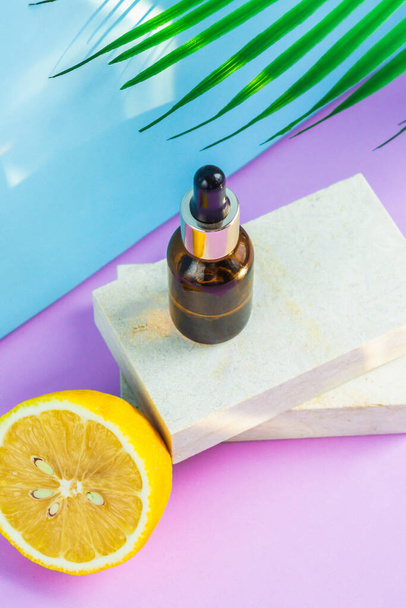 A cosmetic jar with an eyedropper stands on two granite slabs, like on a catwalk, next to half a yellow lemon, a palm leaf and the glare of the sun in the background. Cosmetics for summer. - Foto, Bild