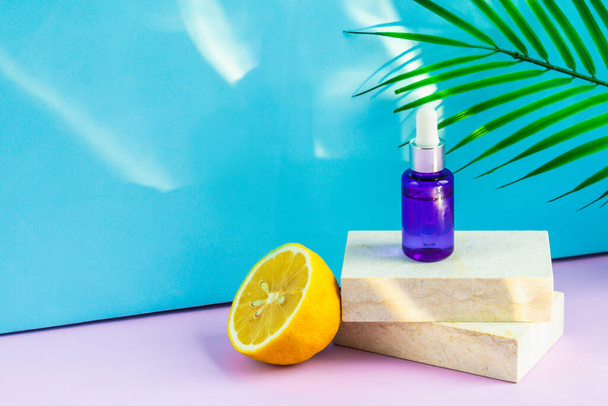A cosmetic jar with an eyedropper stands on two granite slabs, like on a catwalk, next to half a yellow lemon, a palm leaf and the glare of the sun in the background. Cosmetics for summer. - Foto, immagini