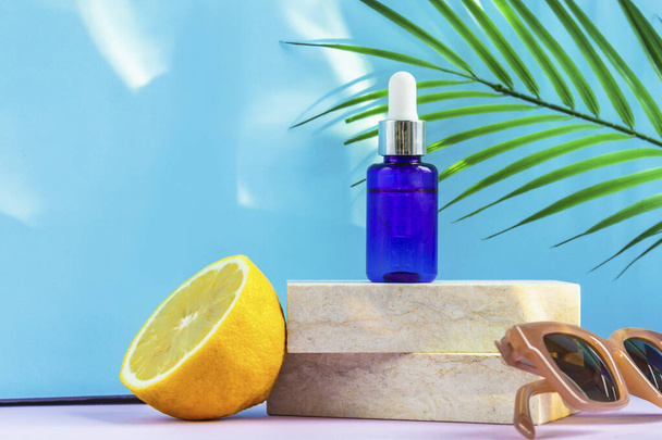 A cosmetic jar with an eyedropper stands on two granite slabs, like on a catwalk, next to half a yellow lemon, a palm leaf, sunglasses and the glare of the sun in the background. Cosmetics for summer. - Photo, Image