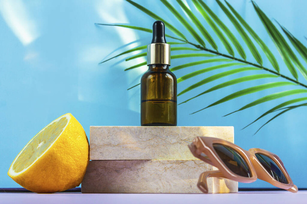 A cosmetic jar with an eyedropper stands on two granite slabs, like on a catwalk, next to half a yellow lemon, a palm leaf, sunglasses and the glare of the sun in the background. Cosmetics for summer. - Photo, image