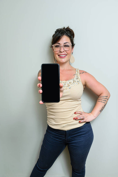 Бразильская женщина в очках, с татуировками, волосы вверх, улыбаясь и показывая свой смартфон в камеру. - Фото, изображение