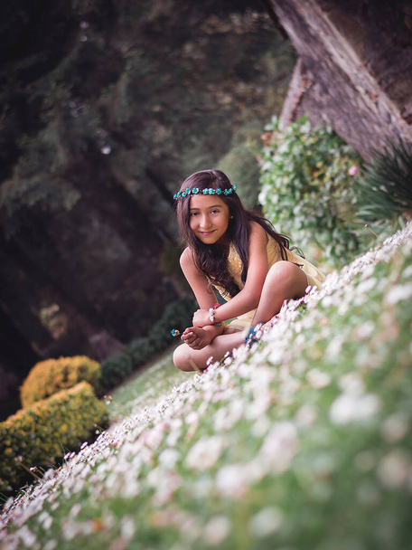 Nastolatka siedząca na trawie w parku w żółtej sukience hipisowskiej i koronie kwiatowej uśmiechnięta do aparatu - Zdjęcie, obraz
