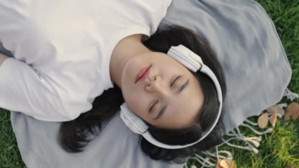 Mujer asiática que usa auriculares escuchando música mientras está tumbada en el suelo de hierba al aire libre en un parque público en la hermosa puesta de sol. Sensación de libertad relajante concepto de estilo de vida. - Imágenes, Vídeo