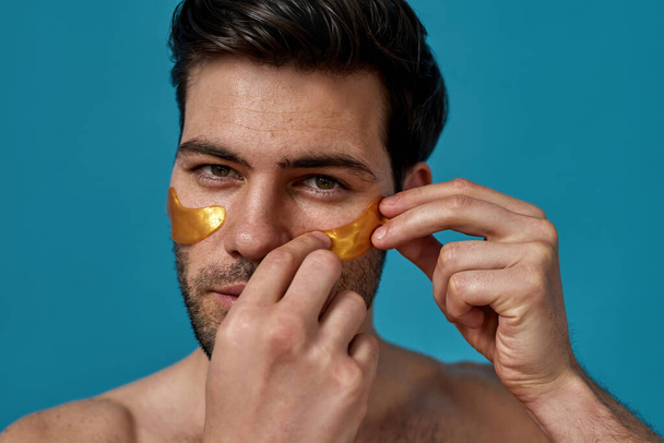 Handsome brunette man applying golden gel patches under his eyes, looking at camera, posing isolated over blue background - Foto, Imagem