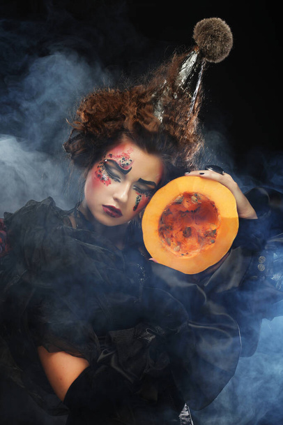Halloween witch with pumpkin, young woman with bright make up and creative hairstyle over dark background - Photo, image