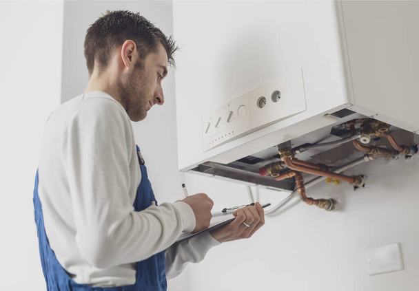 Professional plumber servicing a boiler and writing on a clipboard - Photo, image