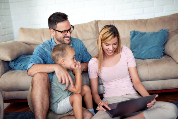 Rodzina korzystająca z laptopa podczas siedzenia na podłodze w salonie, mają wideo połączenie z przyjaciółmi. - Zdjęcie, obraz
