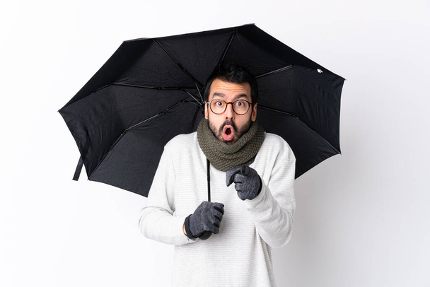 Caucásico guapo hombre con barba sosteniendo un paraguas sobre aislada pared blanca sorprendido y apuntando hacia delante - Foto, imagen
