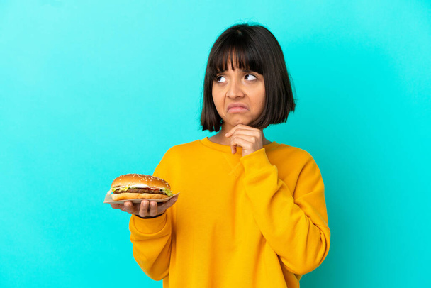Junge brünette Frau hält einen Burger über isoliertem Hintergrund und hat Zweifel - Foto, Bild