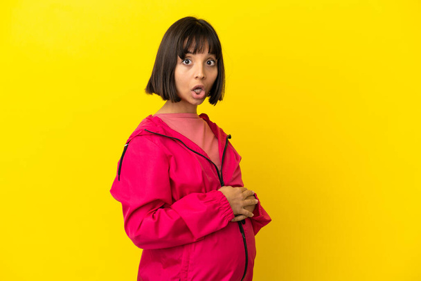 Yüzünde sürpriz bir ifade olan, izole edilmiş sarı arka planda genç hamile bir kadın. - Fotoğraf, Görsel