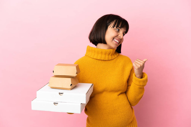 Pembe arka planda pizza ve hamburger tutan hamile bir kadın bir ürün sunmak için kenarı işaret ediyor. - Fotoğraf, Görsel