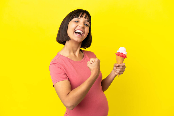Έγκυος γυναίκα κρατώντας ένα παγωτό κορνέτα απομονώνονται σε κίτρινο φόντο γιορτάζει μια νίκη - Φωτογραφία, εικόνα