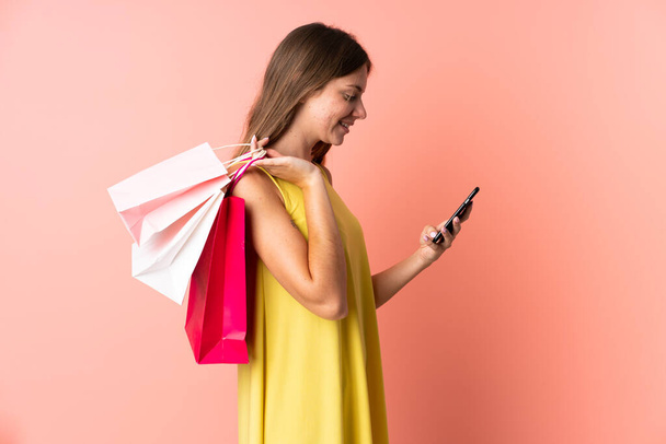 Giovane donna lituana isolata su sfondo rosa che tiene borse della spesa e scrive un messaggio con il suo cellulare ad un amico - Foto, immagini