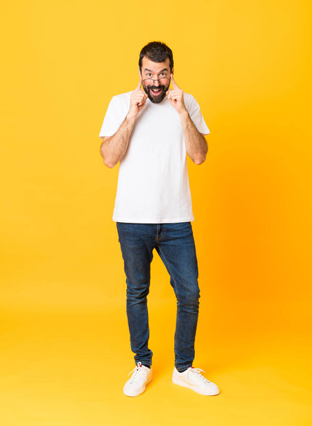 Full-length shot van de mens met baard over geïsoleerde gele achtergrond met bril en verrast - Foto, afbeelding