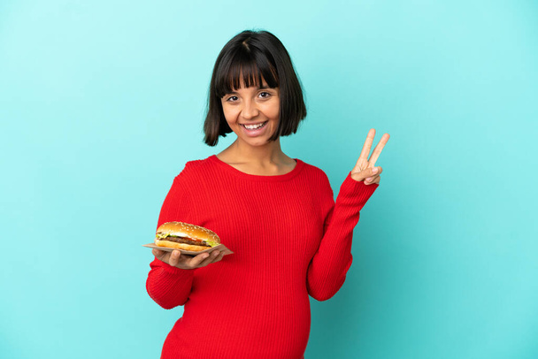 Młoda ciężarna kobieta trzyma hamburgera na odosobnionym tle uśmiechając się i pokazując znak zwycięstwa - Zdjęcie, obraz